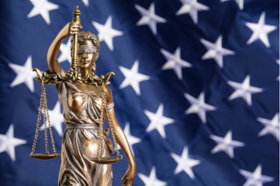 Bronze statue of Lady Justice holding scales in front of an American flag with white stars.